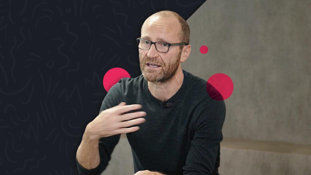 A man with glasses and a beard gestures while speaking, wearing a black sweater. The background is dark with abstract red shapes.