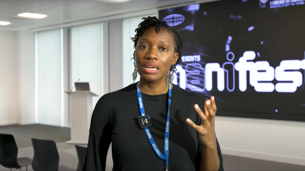 A person speaking passionately in a modern conference room, wearing a lanyard and headset. A large screen displays graphics in the background.