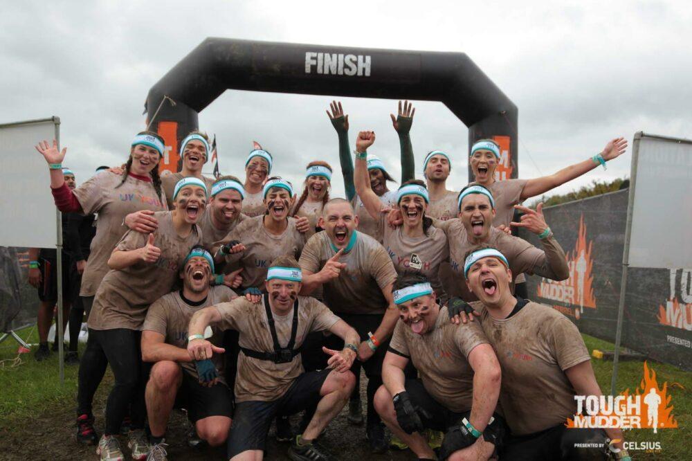 Group of muddy participants celebrating at the finish line of a Tough Mudder event, wearing headbands and raising their arms in victory.