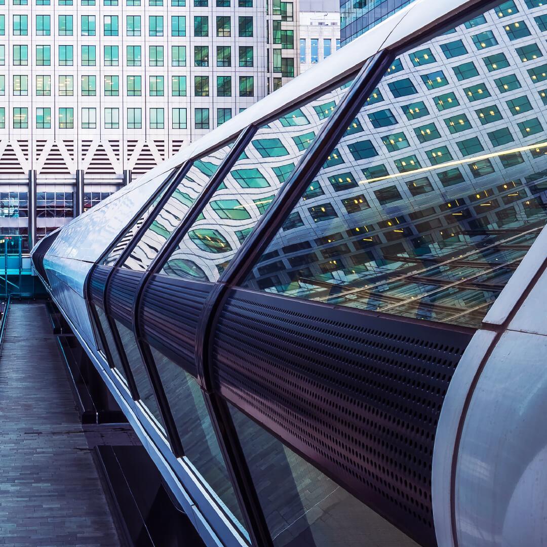 Curved tubular glass walkway linking office towers, reflecting a grid of windows in its mirrored panes.