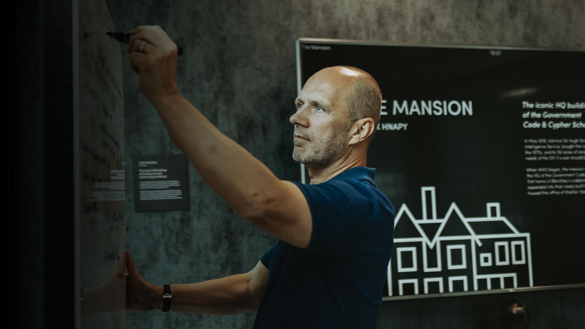 Man writing on a glass board in a dimly lit room with a sign in the background displaying "The Mansion."