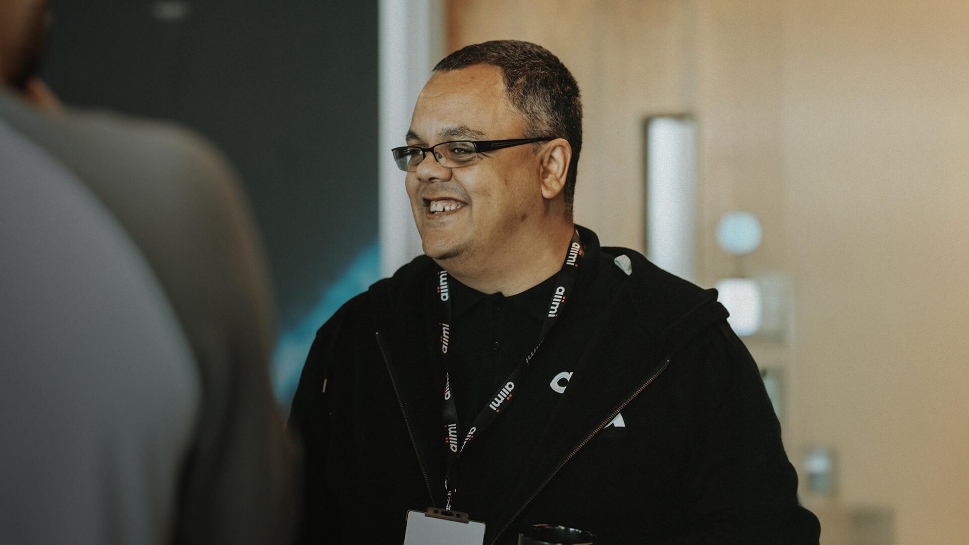 Smiling person wearing glasses and a black hoodie with a lanyard, standing indoors.