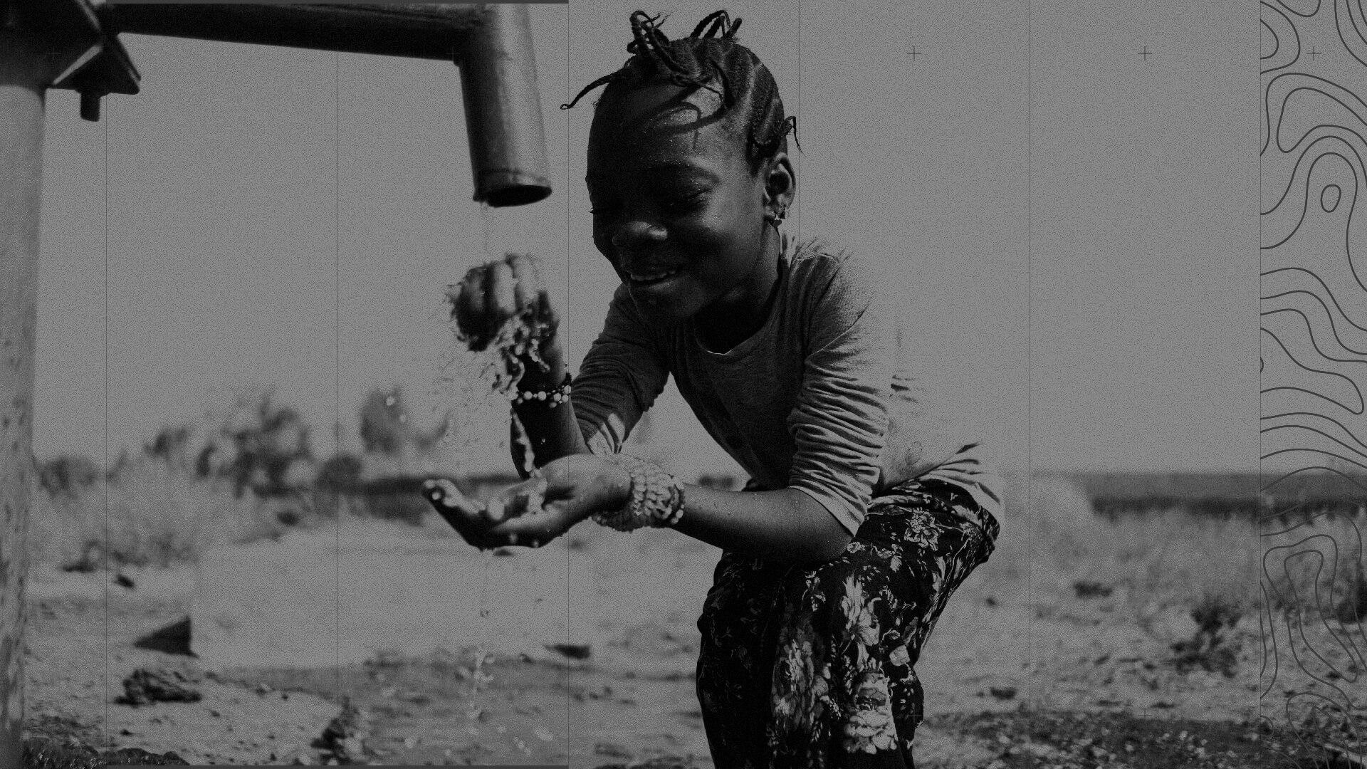 A child joyfully collects water from a pump outdoors, with water splashing into cupped hands. The scene is in black and white.