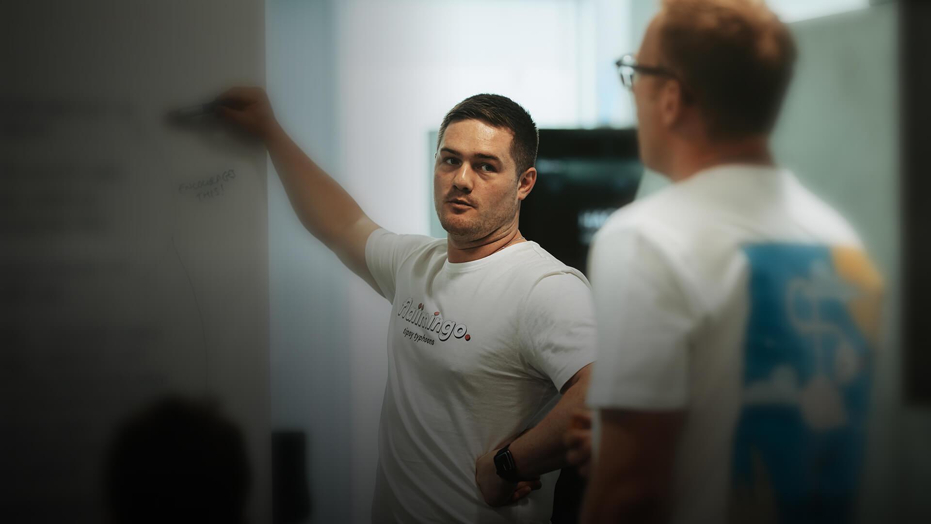 Two people in a meeting room, one writing on a whiteboard while the other listens attentively. Both are wearing casual t-shirts.