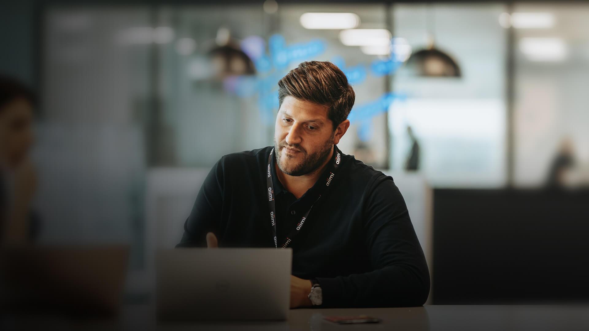 Man with a lanyard working on a laptop in a modern office setting, with blurred background and pendant lights.
