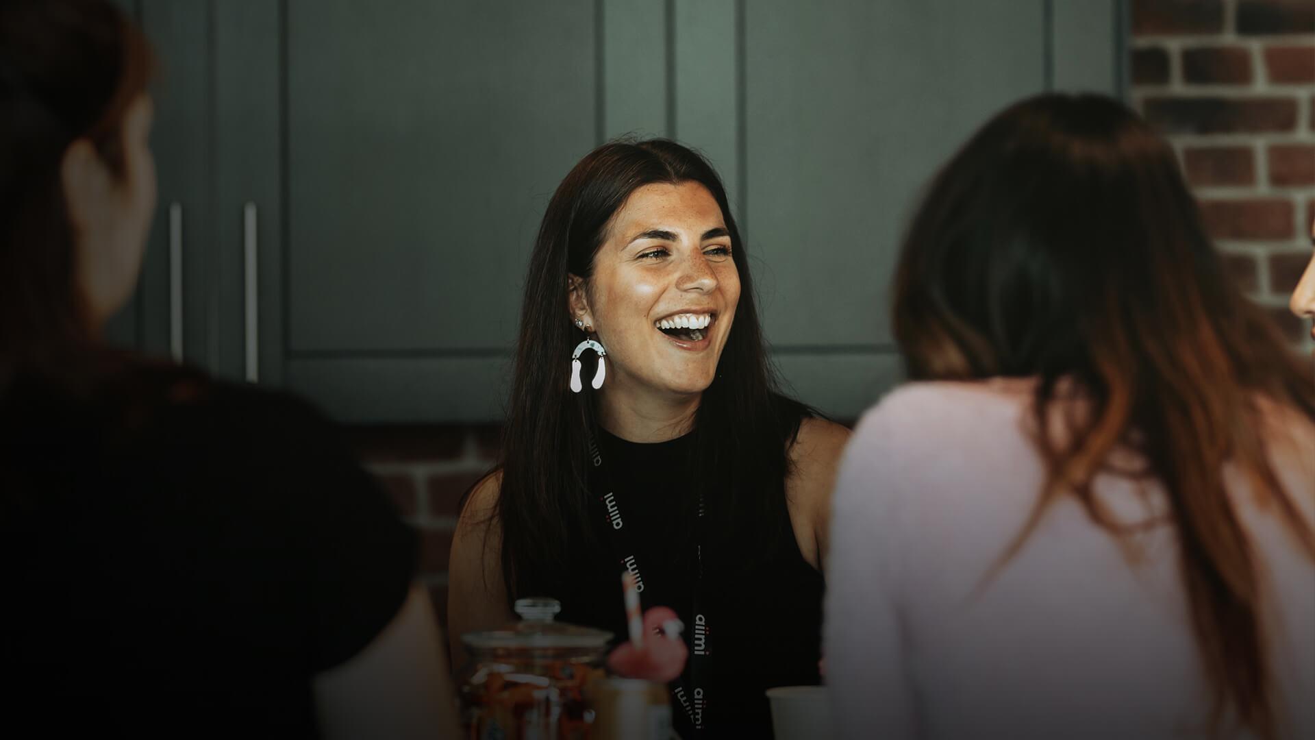 Three people are sitting and talking in a cozy room with brick walls. One woman in the center is laughing, creating a lively atmosphere.