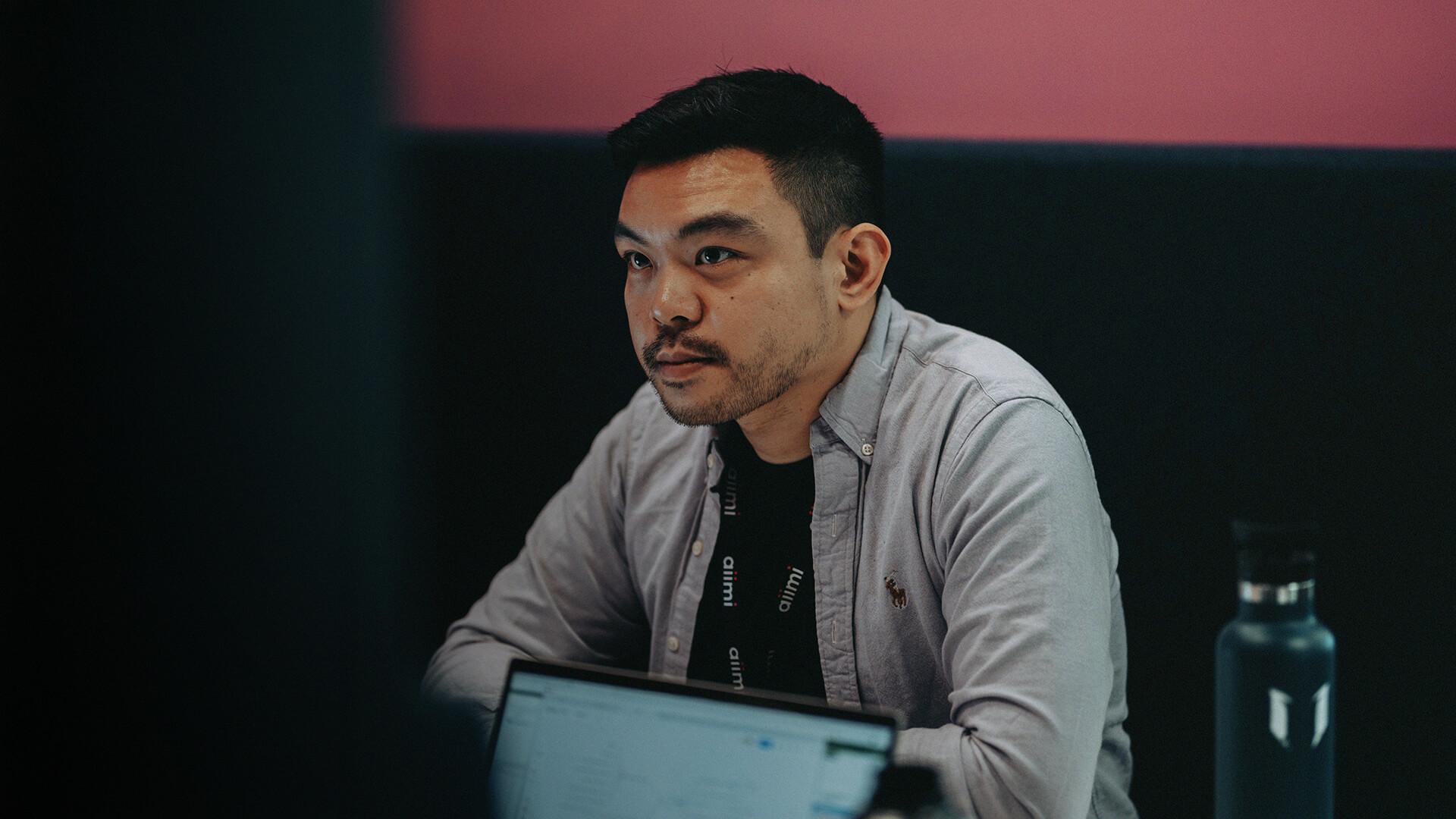 Man focused on a computer screen, wearing a light shirt and lanyard, with a water bottle nearby. Background is dark with a pink wall.