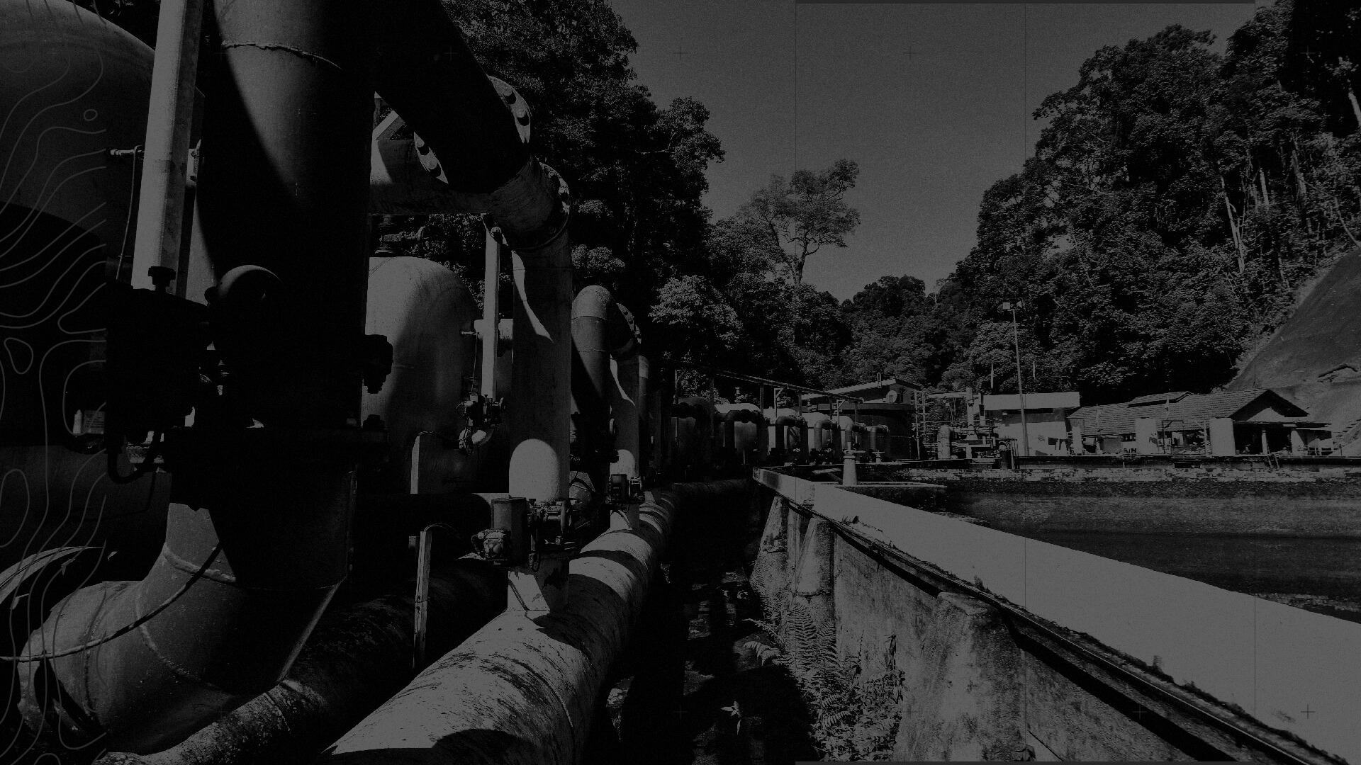 Industrial pipes and machinery in a forested area, with a small building in the background, under a clear sky. Black and white image.