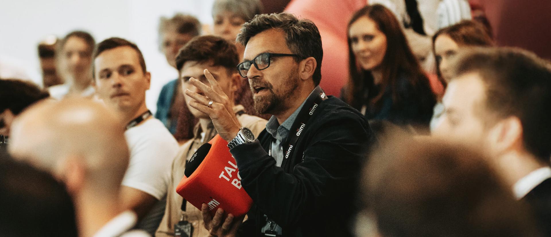 Man wearing glasses gestures while holding a red microphone cube at a crowded conference Q&A.