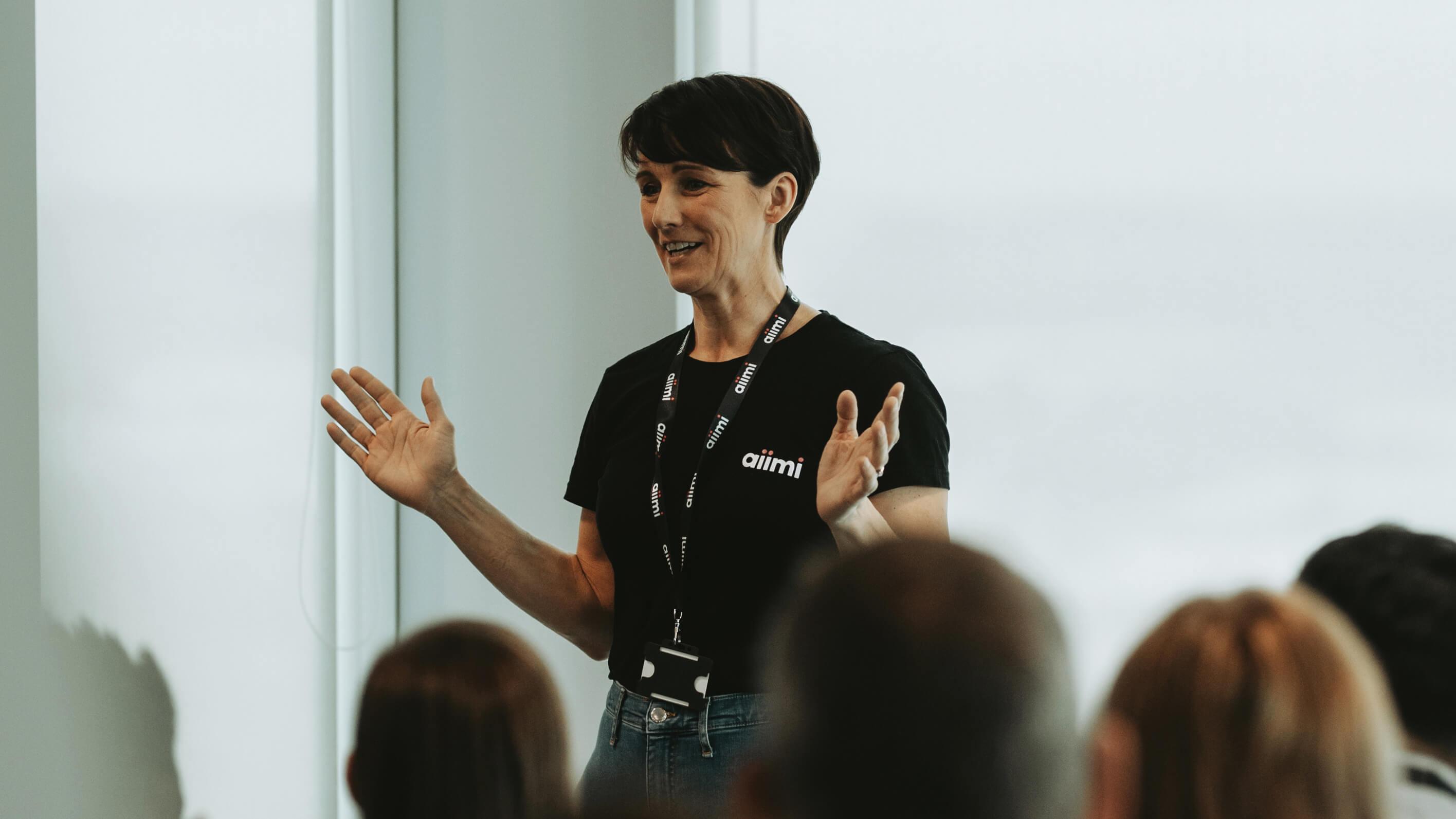Person speaking enthusiastically to an audience, wearing a black "aiimi" shirt and lanyard, gesturing with hands in a bright room.