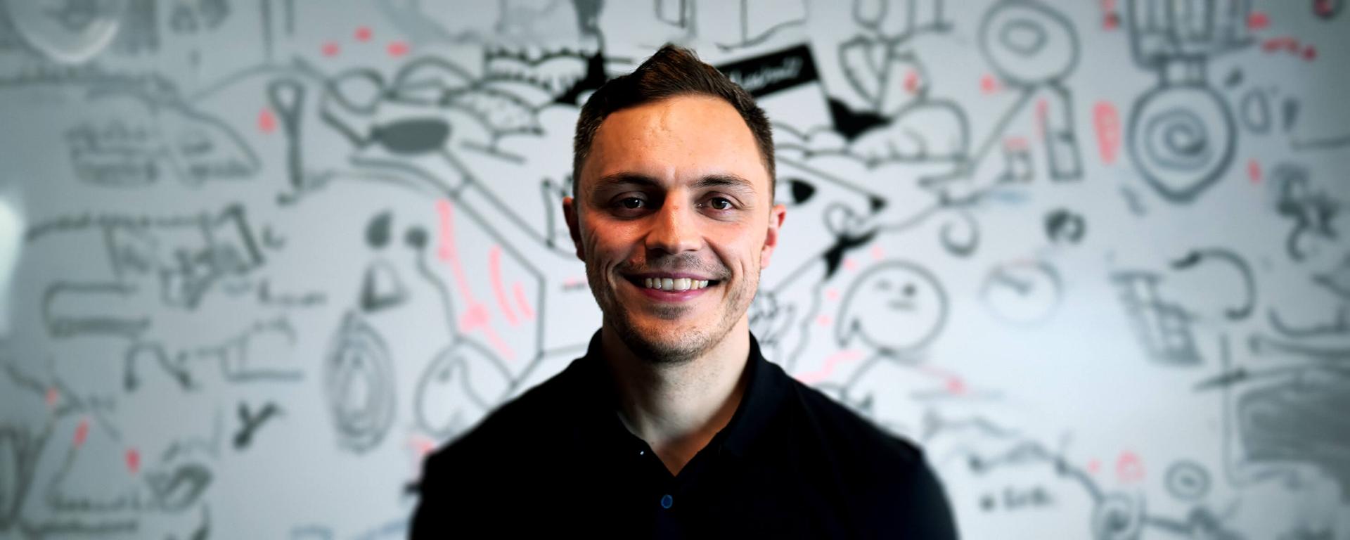 Smiling man in a black shirt stands in front of a whiteboard filled with abstract doodles and sketches.