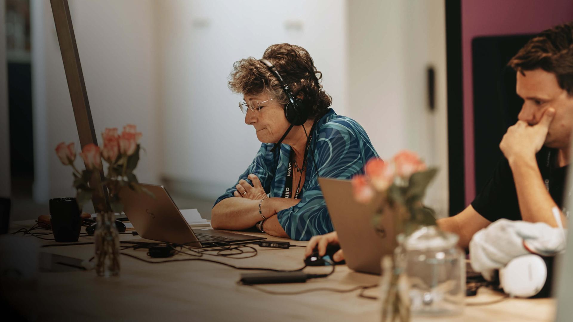 A person wearing headphones sits at a table with a laptop, surrounded by flowers and another person, focused on their work.