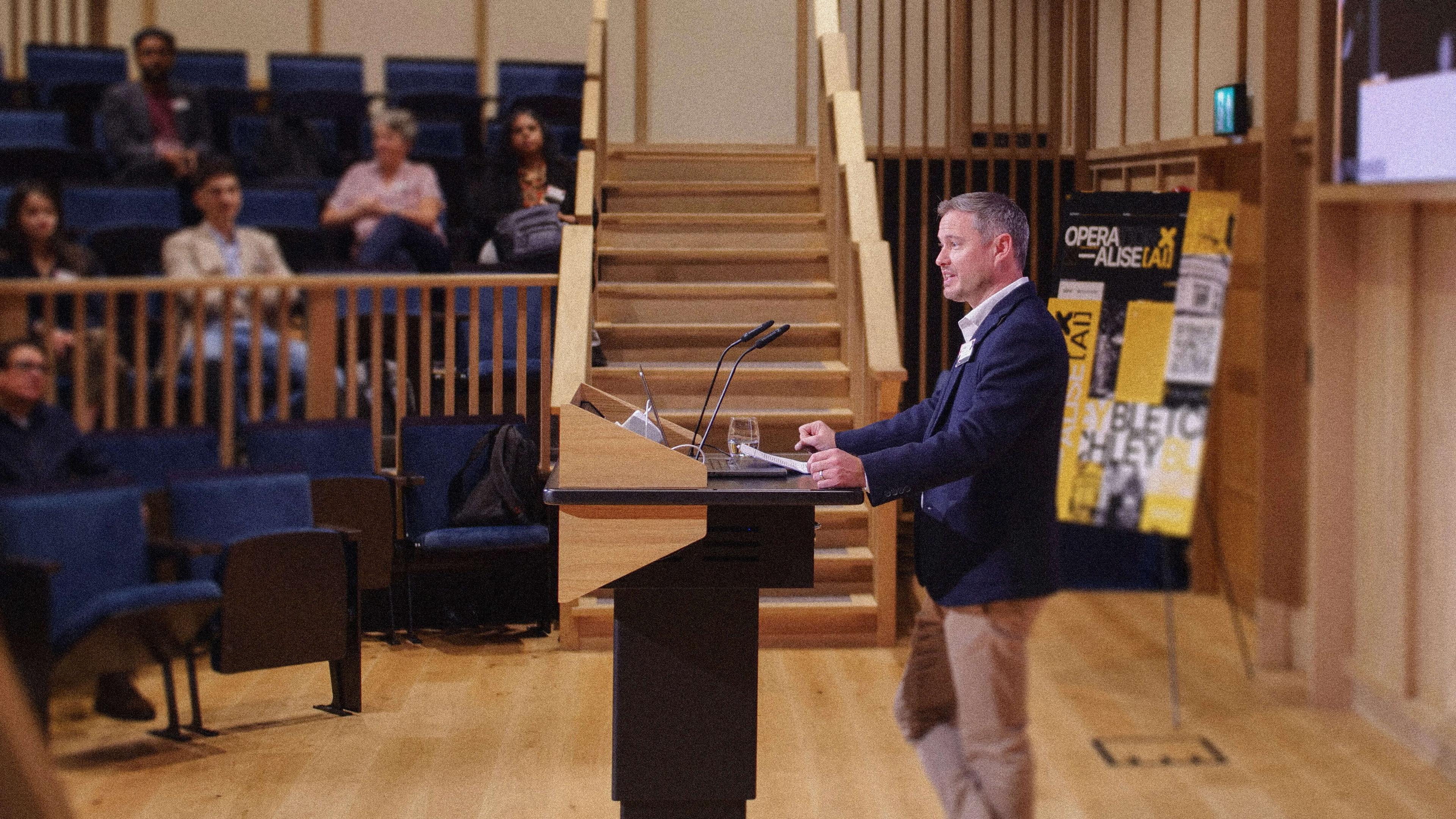 A man in a suit speaks at a podium in a lecture hall, with an audience seated and posters on a stand nearby.