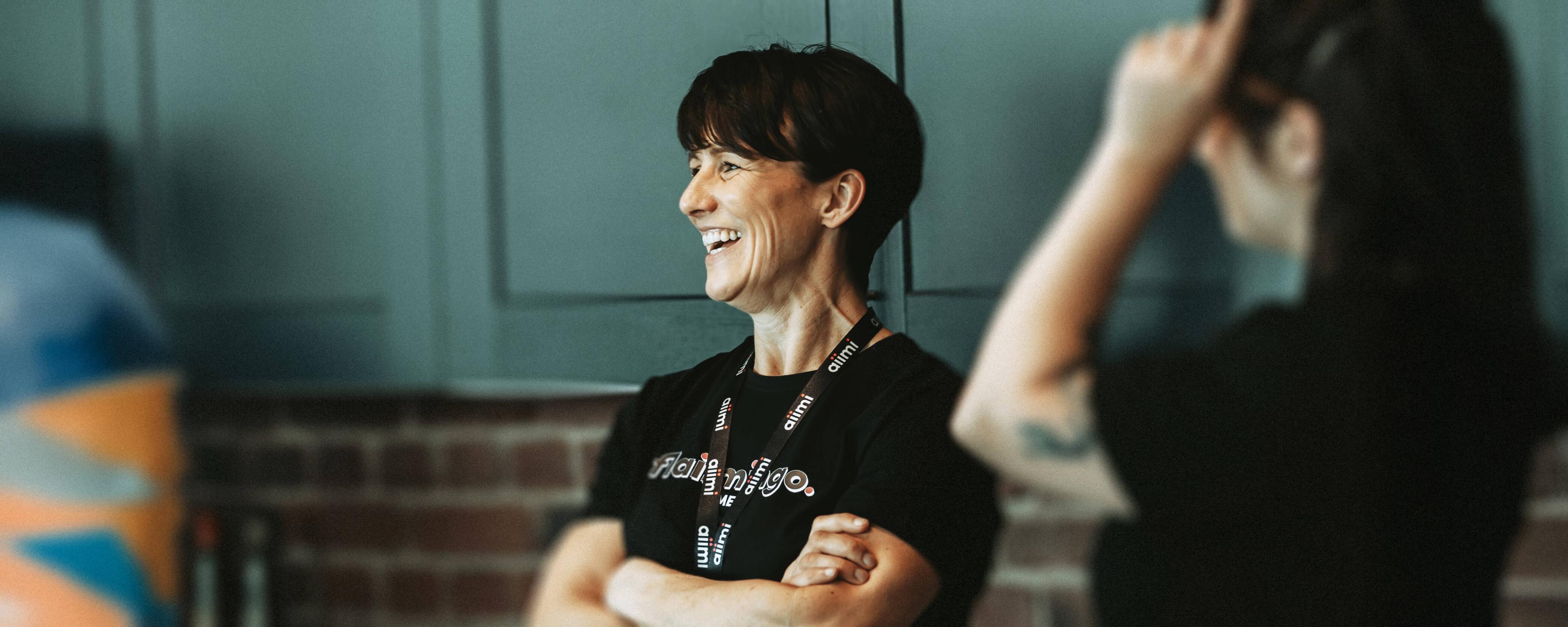 Smiling woman with short hair and a lanyard stands with arms crossed, engaging in conversation. Another person is partially visible beside her.