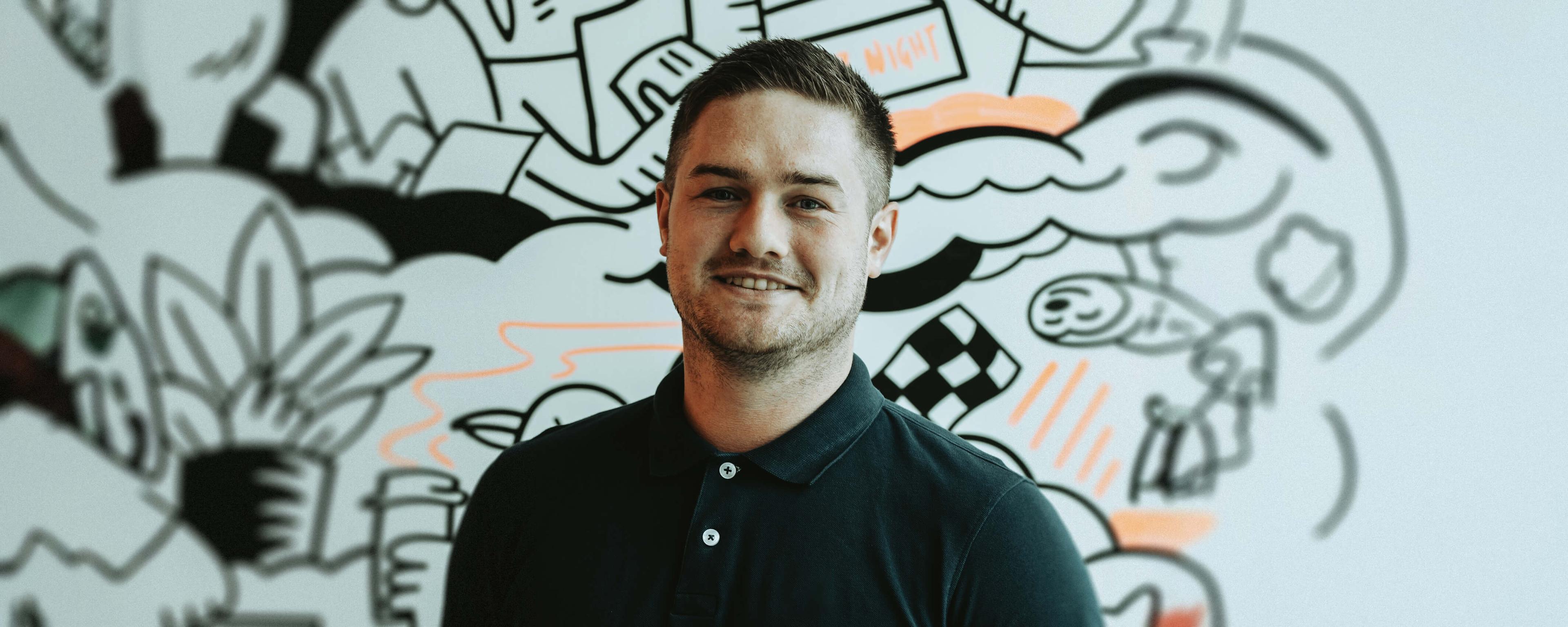 Smiling man in a black polo shirt stands in front of a colorful, abstract mural featuring various doodles and shapes.