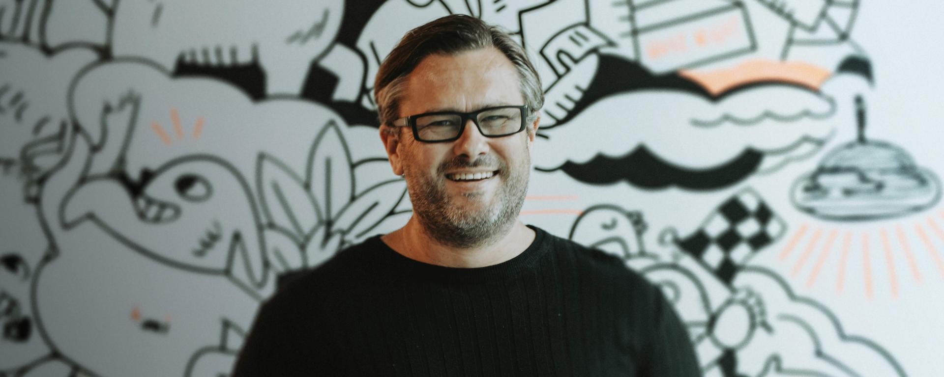 A person wearing glasses and a black sweater smiles in front of a whimsical, black-and-white mural with various doodles and patterns.