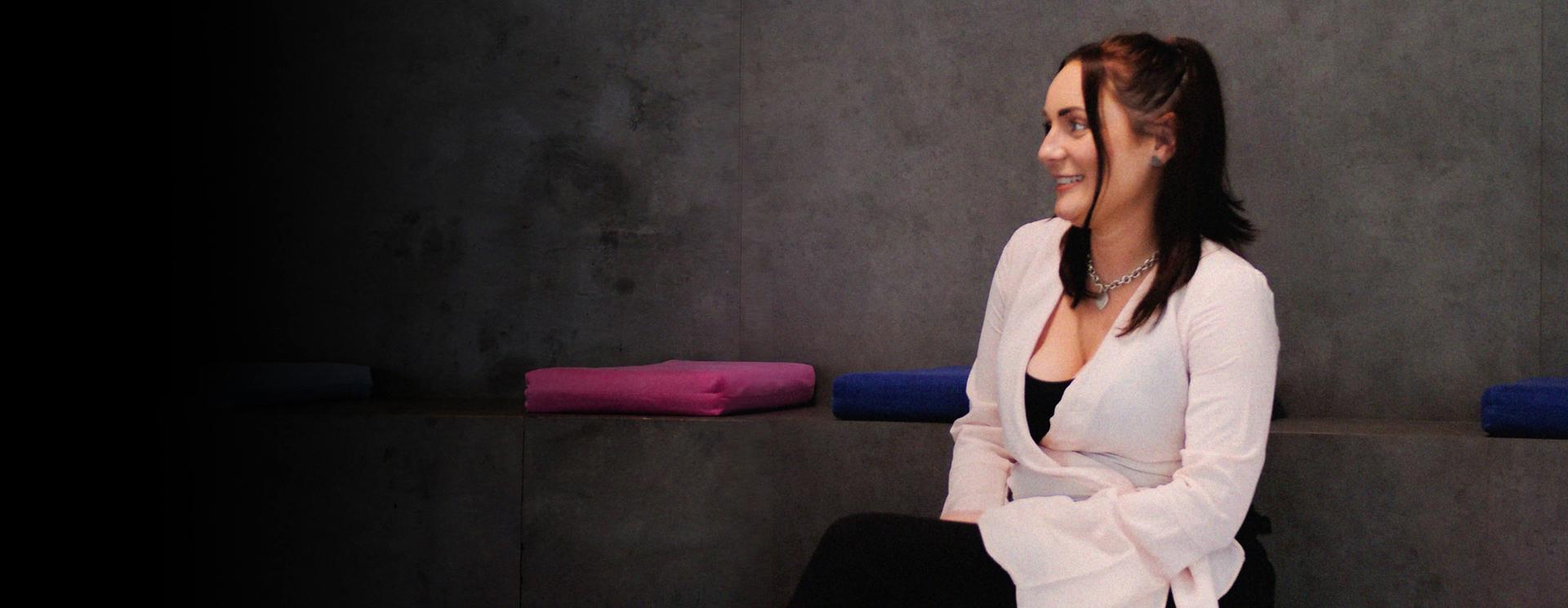 A smiling woman with dark hair sits on a bench with colorful cushions against a gray wall.