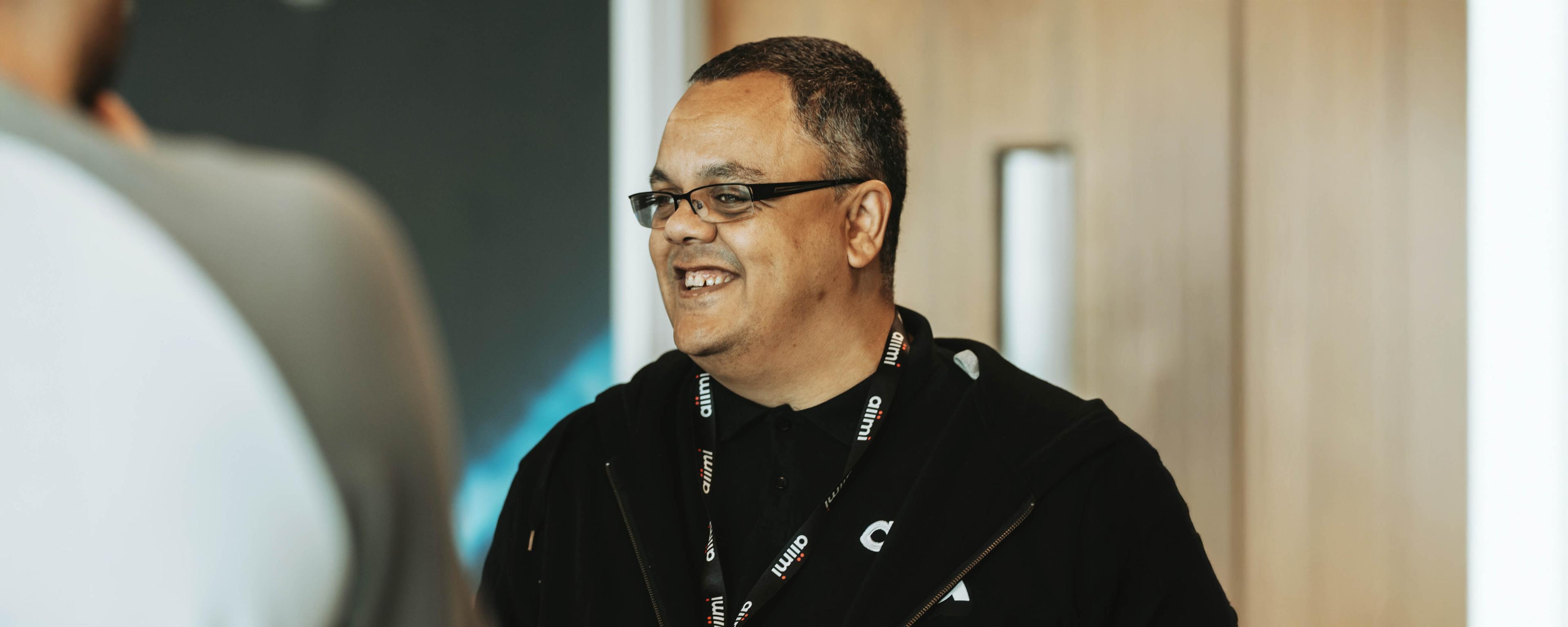 Smiling person wearing glasses and a lanyard, standing indoors with a blurred background.
