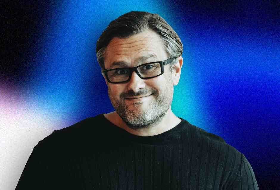 Man with glasses and a beard, wearing a black sweater, smiling against a blue and purple gradient background.