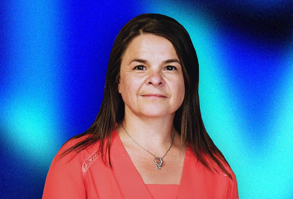 A woman with long brown hair in a red blouse stands against a blue gradient background, wearing a necklace with a small pendant.