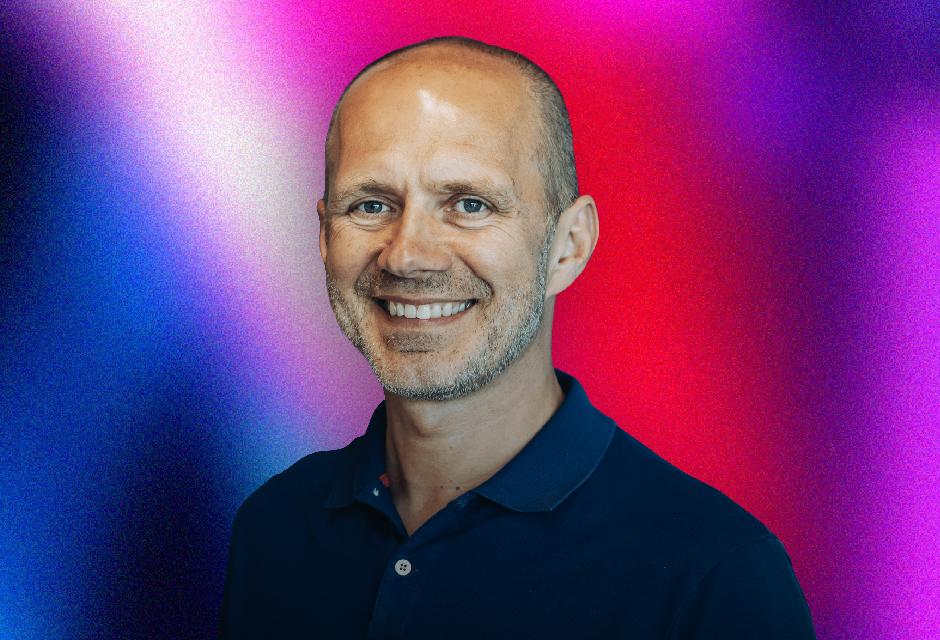 Smiling man in a navy shirt against a vibrant gradient background of blue, pink, and red hues.